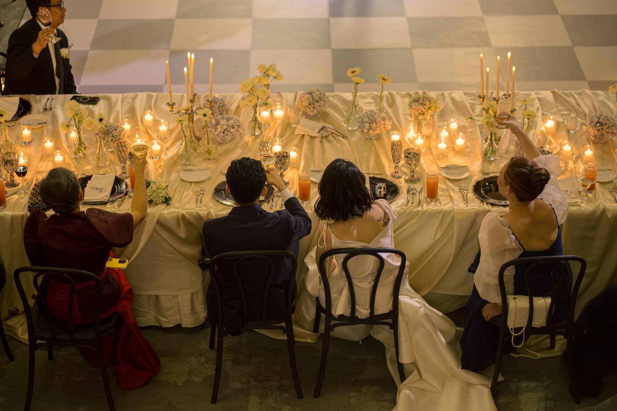 Guests and the newlyweds raising their glasses in a toast along a candlelit head table dressed with pale blooms