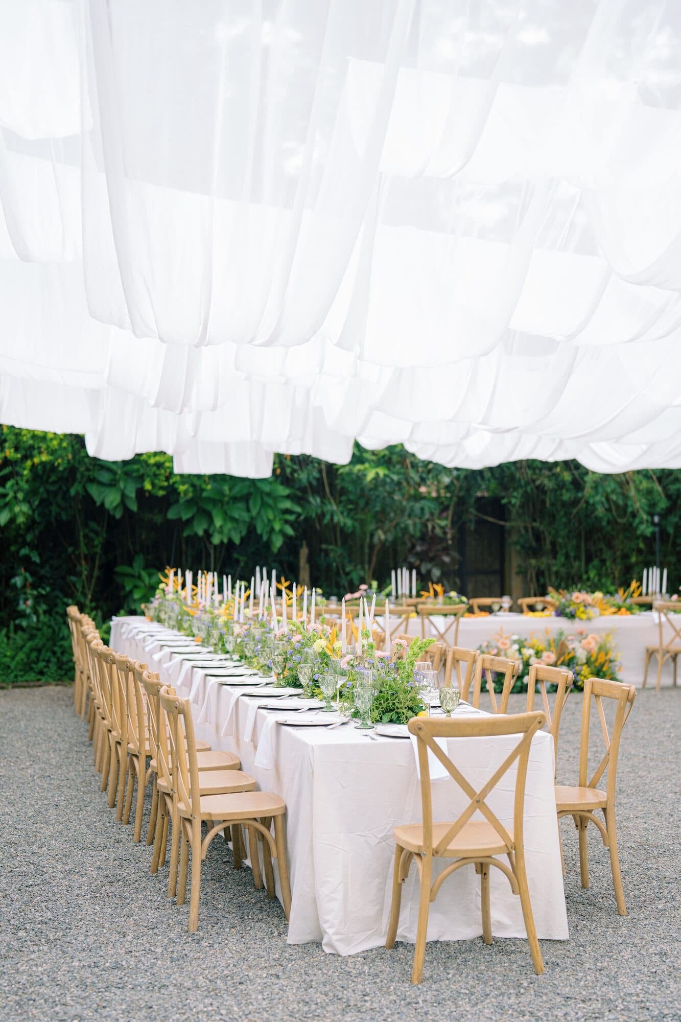 The long reception table under a white fabric canopy, set with candles, greenery and wooden crossback chairs