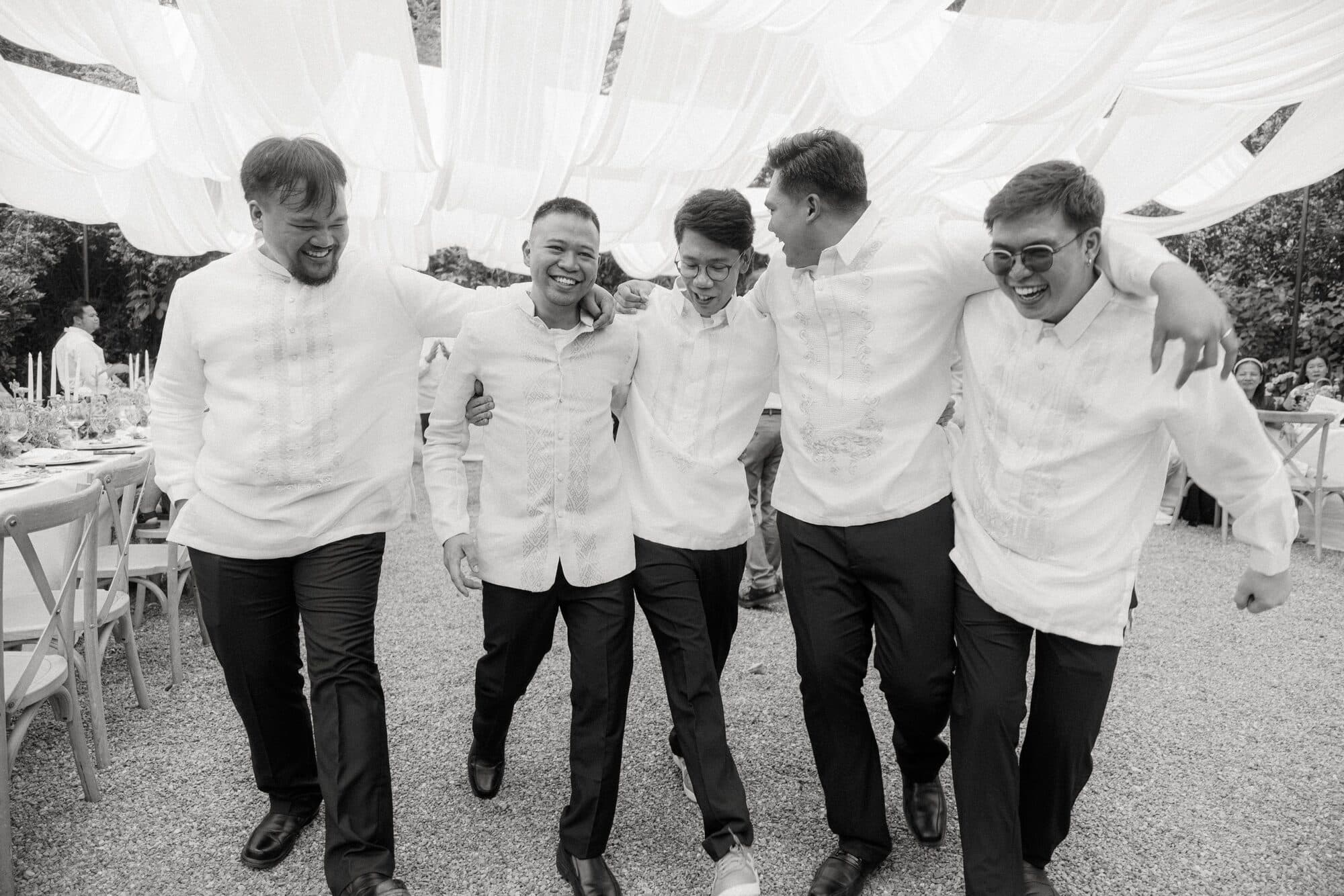 Five groomsmen in barongs walking arm in arm across the garden, laughing
