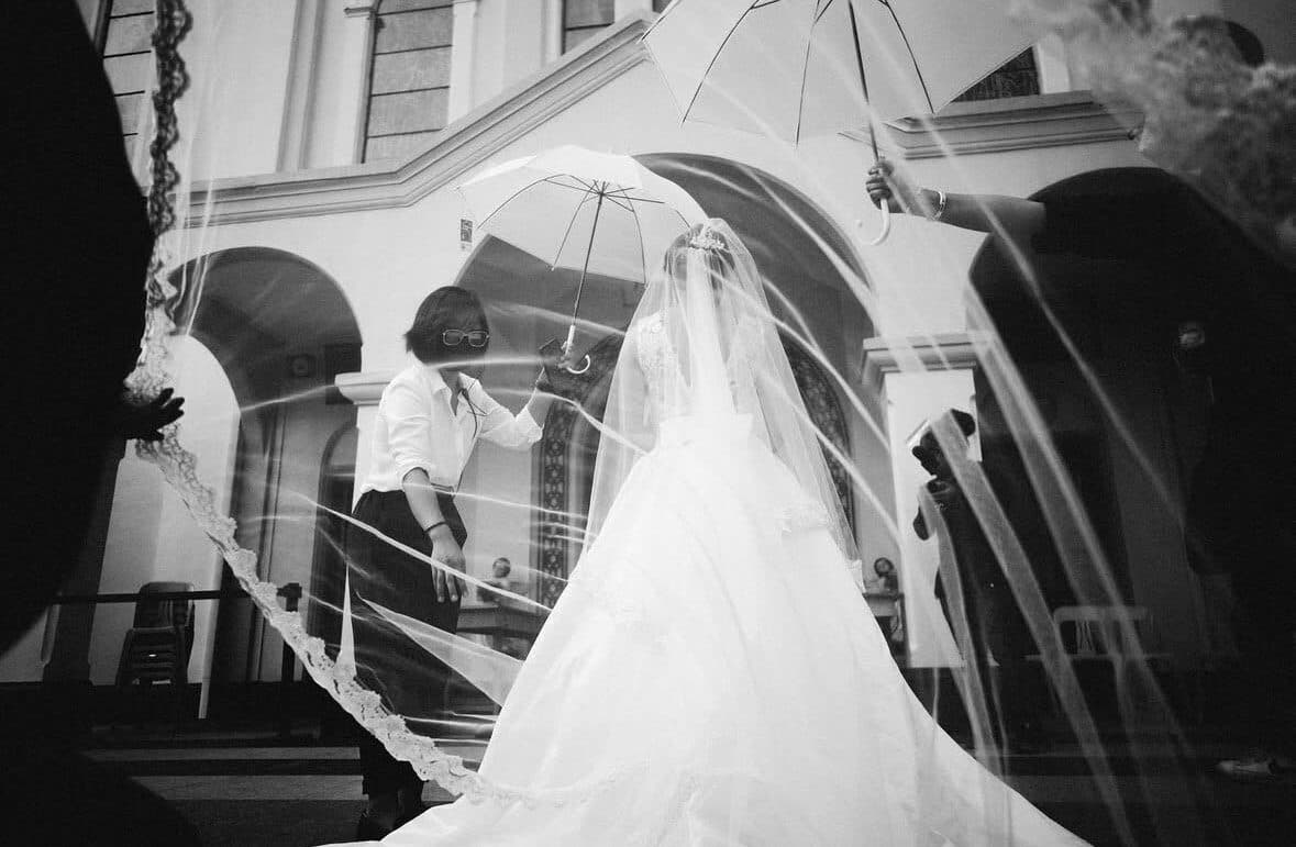 A bride sheltered under a clear umbrella outside a church, her veil lifting in the wind as a coordinator steadies it